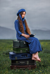 Dreamy vintage girl in blue dress reading a book on stacked suitcases in countryside field under cloudy sky