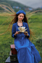 Dreamy vintage girl in blue dress reading a book on stacked suitcases in countryside field under cloudy sky