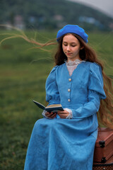 Dreamy vintage girl in blue dress reading a book on stacked suitcases in countryside field under cloudy sky