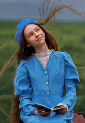 Dreamy vintage girl in blue dress reading a book on stacked suitcases in countryside field under cloudy sky