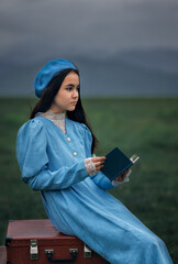 Dreamy vintage girl in blue dress reading a book on stacked suitcases in countryside field under cloudy sky
