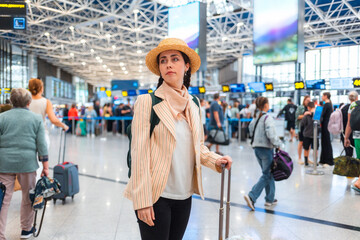 Pretty Caucasian young woman in straw hat wheeling a suitcase to the check-in desk. Defocused people in background. The concept of vacation and flight trip