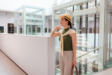 Mid shot of young pretty Caucasian elegant woman in suit and straw hat holds bag and posing in the modern art gallery. Concept of cultural education and Museum day