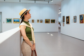 Mid shot of young Caucasian elegant woman in suit and straw hat contemplating pictures in the modern art gallery. Side view. Concept of cultural education and Museum day