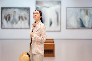 Mid shot of young Caucasian woman in elegant jacket, straw hat and trousers contemplating in the art gallery. Copy space. Concept of cultural education for students and Museum Day