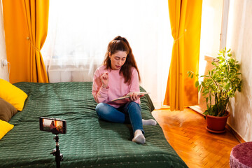 Influencer recording a vlog while sitting on a bed, engaging her audience with inspiring content for digital marketing. Wide shot, top view