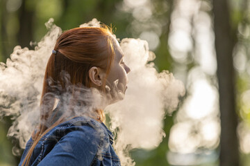 An young woman with long, red hair vaping in a forested area. The person is captured from the side, exhaling a thick cloud of vapor from an electronic cigarette. Summer landscape in a background.