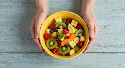 Hands present a vibrant fruit salad bursting with raspberries kiwis and blueberries in a yellow bowl on a rustic wooden table