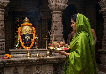 South Asian Woman in Green Silk Saree Carrying Puja Thali to Lord Shiva's Shrine in Intricately Carved Temple