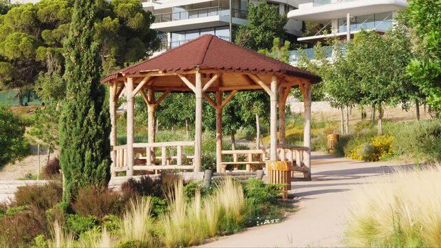 Close-up of a wooden gazebo with brown roof in lush public park in Varkiza, Athens, Greece. Surrounded by green plants, including Nassella tenuissima and Apodasmia similis. No people visible.