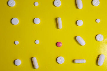 White pills and capsules of various sizes, along with one pink tablet, are arranged in a pattern on a vibrant yellow background.