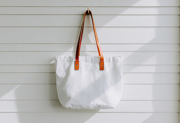 Stylish white tote bag for mockup with brown leather straps hanging against a minimalist white wooden backdrop in bright sunlight