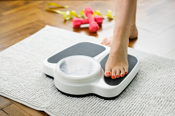 Young woman stepping on scale with dumbbells and measuring tape on bath towel