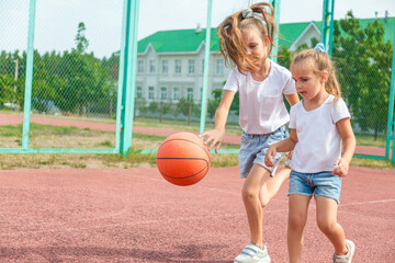 Girl and her younger sister, play basketball on modern basketball court under open sky. Concept of sports, hobbies and healthy lifestyle.