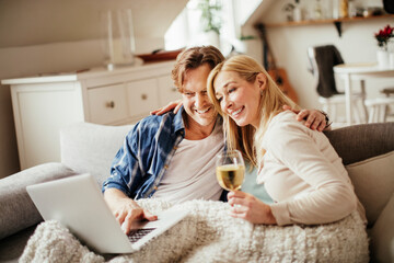 Mature couple having a video call on their laptop while enjoying a glass of wine on the couch in...