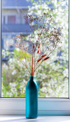 Dried flowers in a beautiful green bottle on the background of a window on a spring day.