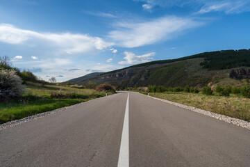 An empty highway that passes through fields and goes towards the mountain. The road is covered with blue sky and beautiful fluffy clouds.	