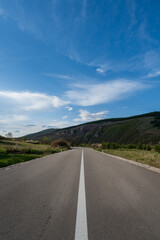 An empty highway that passes through fields and goes towards the mountain. The road is covered with blue sky and beautiful fluffy clouds.	