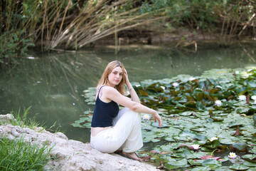 A young, beautiful blonde woman touches her hair and head with one hand, sitting on a rock next to...