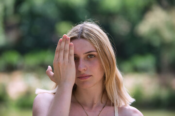 A young, beautiful blonde woman covers half her face with her hand and looks at the camera.