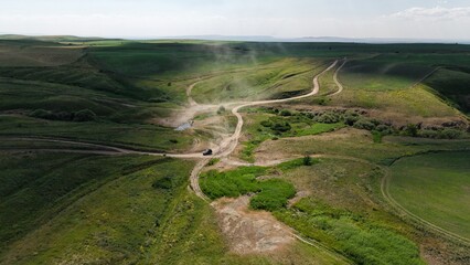 Aerial view of an off road vehicle kicking up dust as it navigates a winding dirt road through rolling green hills, creating a dynamic scene of adventure and exploration in the countryside