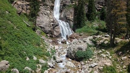 Refreshing waterfall cascading down a steep, rocky mountainside, creating a picturesque scene amidst lush green vegetation and a clear blue sky, evoking tranquility and the beauty of nature