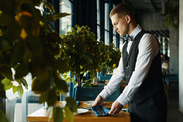 Caucasian young adult man arranging table setting in restaurant, standing beside wooden table with...
