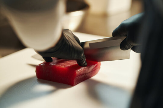 Caucasian young adult man wearing black gloves slicing raw tuna fillet with sharp knife on cutting board in professional kitchen, hands visible preparing fresh seafood for cooking