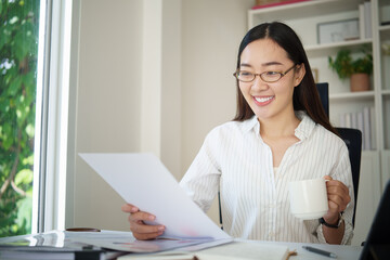 Asian businesswoman reading a report while enjoying a coffee break at her desk.