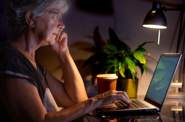 Modern elderly woman freelancer working on computer at night. Attractive businesswoman studying...