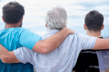 Rear view of a family group of three multigenerational males, father, son and nephew hugging together outdoors by the seaside feeling affection, care, freedom and vacation