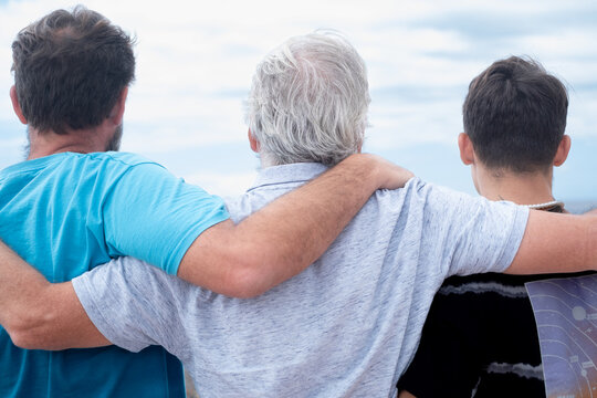 Rear view of a family group of three multigenerational males, father, son and nephew hugging together outdoors by the seaside feeling affection, care, freedom and vacation