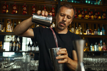 Caucasian middle aged man preparing cocktail behind bar counter, pouring liquid from shaker into mixing tin, concentrating on bartending task with shelves of bottles in background