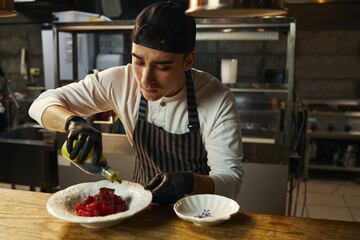 Young adult Caucasian man wearing black gloves preparing gourmet dish in professional kitchen, pouring sauce over diced food on plate, focused on culinary presentation