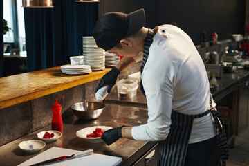 Caucasian young adult man wearing apron and cap preparing dessert in professional kitchen,...