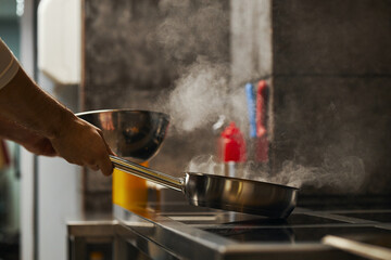 Caucasian middle aged man holding frying pan over stove with steam rising, cooking food in professional kitchen, partial view of arm and hand visible, stainless steel bowl nearby