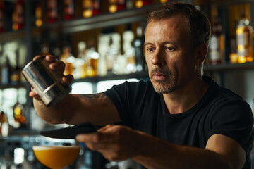 Middle aged Caucasian man preparing cocktail behind bar counter, holding shaker and strainer while pouring drink into glass, focused expression, bottles visible in background