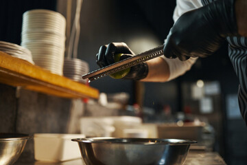 Caucasian young adult man grating lime over metal bowl in professional kitchen, wearing black...
