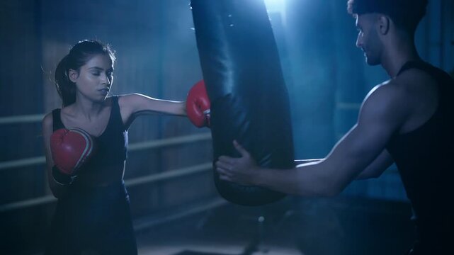 Indian asian young adult gen z angry woman teen age girl do sport training indoor place athlete fit male trainer boxer teach skill beat boxing bag female lady wear gloves fist hand in dark class room