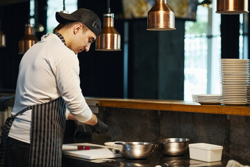 Young adult Caucasian man wearing apron preparing food in professional kitchen, standing at counter slicing ingredients with knife, stacks of plates and metal bowls nearby