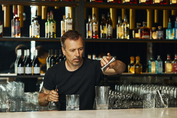 Middle aged Caucasian man preparing cocktail behind bar counter, holding shaker and pouring drink into glass, surrounded by bottles and glassware, focused on bartending
