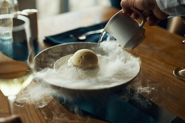 Caucasian male hand pouring liquid from small pitcher onto dessert in bowl with dramatic vapor...