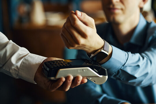 Caucasian young adult man paying with smartwatch by holding wrist near payment terminal, hand of another holding device, focus on contactless transaction technology