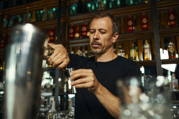 Middle aged Caucasian man preparing cocktail behind bar counter, holding jigger and pouring liquid with focused expression, shelves with bottles visible in background
