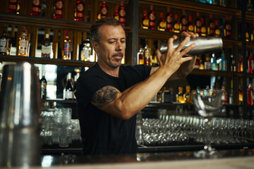 Caucasian middle aged man preparing cocktail behind bar counter, holding shaker with both hands, concentrating on mixing drink, standing in front of shelves with bottles and glassware