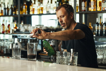 Caucasian middle aged man preparing cocktail behind bar counter, pouring liquid from green bottle into mixing glass, focused on bartending task in modern bar setting