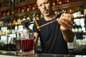 Middle aged Caucasian man preparing cocktail using torch and cinnamon stick in bar setting,...