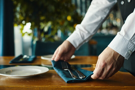 Caucasian young adult man arranging cutlery and napkin on restaurant table, hands visible placing utensils beside empty plates, preparing dining setup for guests - Powered by Adobe