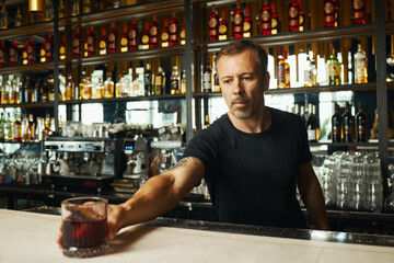 Caucasian middle aged man serving drink at bar counter, reaching forward with glass in hand, standing in front of shelves filled with bottles and glassware, focused expression