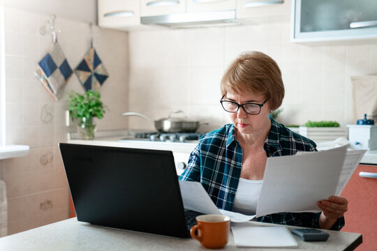 Senior woman working from home kitchen with laptop, documents, finances. Personal budgeting, financial literacy, pension plan, retirement lifestyle. Stress, money anxiety. Ageism in looking for job